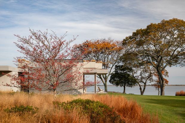 A modern, low-profile waterfront residence viewed from a landscaped garden. The house features pale rendered walls, extensive vertical slatted screening, a flat roof, and a covered outdoor terrace or balcony. Ornamental grasses, autumnal trees, and a manicured lawn frame the building, with calm water visible beyond.