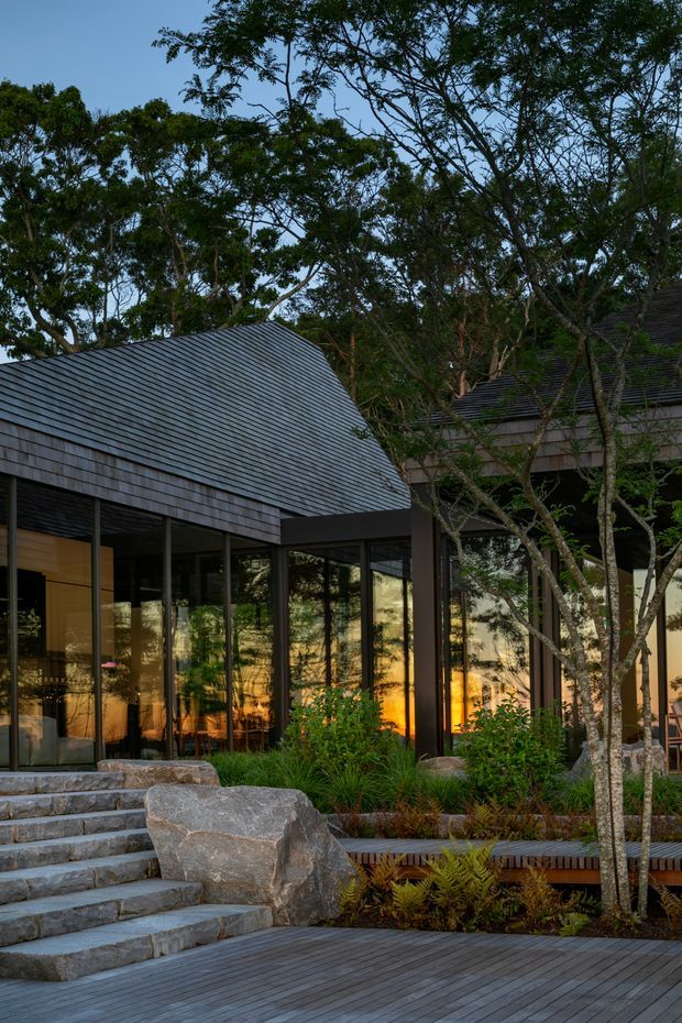 Exterior architectural photograph of a contemporary pavilion-like house set within mature trees and landscaped gardens. The building features steep pitched roof planes clad in dark small-format shingles or tiles, extensive floor-to-ceiling glazing with dark frames, timber decking, broad stone steps, large natural boulders, and integrated planting. Warm interior lighting glows through the glass at dusk.