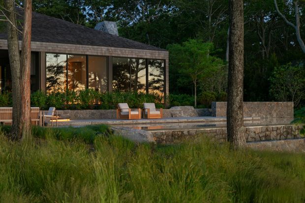 A low, modern pavilion-like house is nestled within a wooded landscape. The dark pitched roof and broad floor-to-ceiling glazed wall frame a warm interior, while a stone terrace with outdoor lounge chairs, built-in stone walls, and subtle lighting extends into the surrounding grasses and trees.