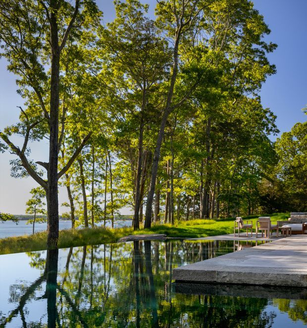 A tranquil landscaped outdoor setting with a dark reflecting swimming pool bordered by mature trees and a broad pale stone or concrete terrace. Outdoor lounge furniture sits beside the pool, while a calm body of water is visible beyond the tree line. The scene is photographed in bright natural daylight and emphasises the pool, terrace, and wooded waterfront landscape.