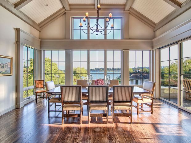 A bright, spacious dining room with a large rectangular dining table and wooden dining chairs arranged in front of expansive floor-to-ceiling windows. The room features warm dark timber flooring, light painted walls, extensive white-framed glazing, a high vaulted shiplap ceiling with exposed beam detailing, and a central traditional chandelier. Water and neighbouring rooftops are visible beyond the windows, while built-in architectural trim and a glazed corner create an airy, elegant atmosphere.