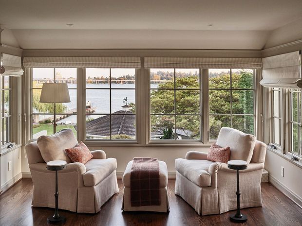 A bright, traditionally styled sitting room with two slipcovered armchairs facing each other across a central ottoman, with small black side tables and a floor lamp. The room has extensive multi-pane corner windows with woven Roman blinds, detailed wall and ceiling trim, and dark polished timber flooring. A waterfront view is visible through the windows.