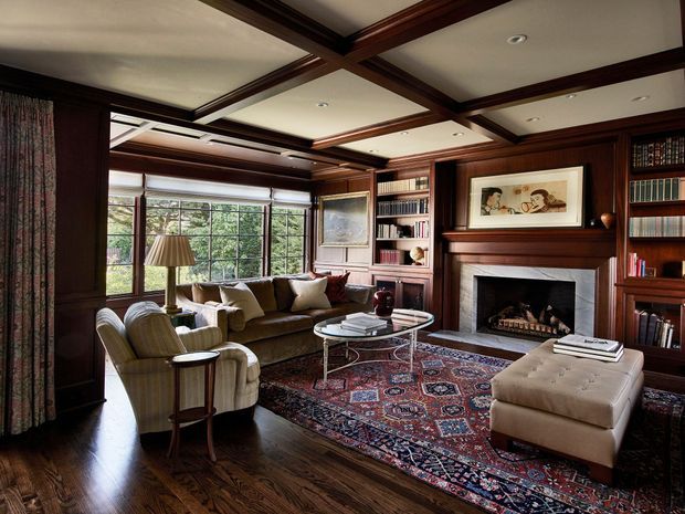 A richly detailed traditional library-style living room with dark timber wall panelling, extensive built-in bookcases, a coffered timber ceiling, and a substantial fireplace with mantel. The room includes upholstered seating arranged around a glass coffee table, a large patterned Persian-style rug, ottomans, and broad windows overlooking greenery. The overall atmosphere is warm, refined, and cosy.