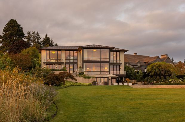 Exterior architectural photograph of a contemporary multi-storey residence set within a landscaped suburban garden. The home has extensive dark-framed floor-to-ceiling glazing, projecting rectangular volumes, low-pitched metal roofs, concrete lower-level walls, and warm interior lighting visible through the windows. Mature trees, ornamental planting, and a broad lawn surround the house, with neighbouring pitched-roof homes in the background.