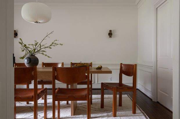 A refined dining room with a rectangular timber dining table and matching mid-century-style wooden chairs upholstered in warm brown leather. The room features white paneled walls and doors, dark timber flooring, a light neutral rug, a sculptural white pendant light, and restrained wall sconces. A ceramic vase with branches and a small centerpiece add natural accents. The overall composition is calm, warm, and understated.