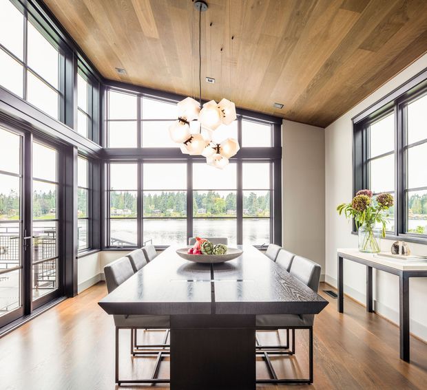 Bright contemporary dining room with a large dark dining table and upholstered chairs, surrounded by expansive dark-framed windows overlooking a waterfront landscape. The room features a warm timber-clad ceiling, timber flooring, minimalist white walls, a sculptural clustered pendant light, and a slim console table with floral arrangement.