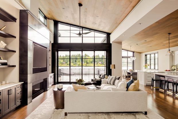 Open-plan living room with a large sectional sofa facing a built-in dark feature wall and fireplace, connected to a bright kitchen and dining area. The space has a double-height glazed opening to a balcony, exposed light timber ceiling, dark-framed windows and doors, hardwood flooring, recessed lighting, and contemporary built-in joinery.
