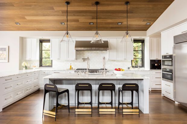 A bright, spacious kitchen with white shaker-style cabinetry, a large central island, integrated appliances, and four dark counter stools. The island and worktops are light-colored, with a white subway-tile backsplash and a stainless-steel range hood. A warm timber ceiling with recessed lights adds contrast, while three clear glass pendant lights hang above the island. Dark-framed windows bring natural light into the room, and the floor has a medium-toned timber finish.