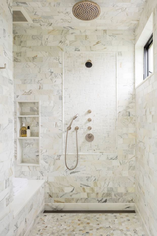 Bright, compact walk-in shower enclosure clad throughout in white and grey veined marble-look tiles. The shower features a large round overhead rain shower, handheld shower, multiple wall controls, a recessed shelf, a small high window, and a pebble-pattern tiled floor. Brushed brass or bronze fixtures add a subtle warm accent.