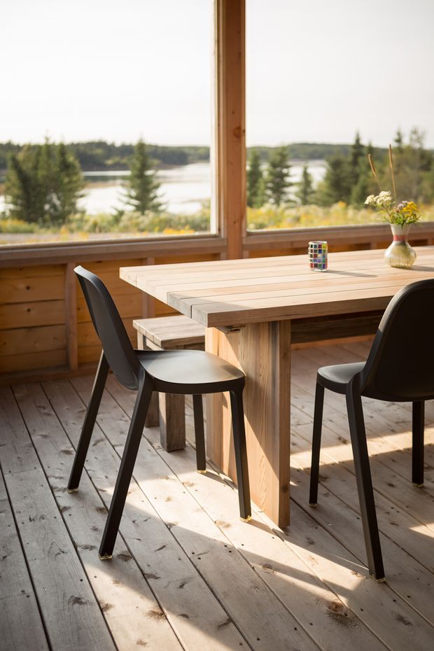 A sunlit dining area in a timber-enclosed room with large windows overlooking a lake and tree-lined landscape. A light timber plank dining table is paired with black dining chairs, with timber wall panelling and wide timber floorboards creating a warm, rustic setting. A small vase of flowers and colourful glass sit on the table.