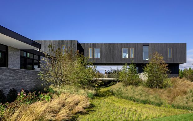 Exterior view of a contemporary multi-level residence composed of dark vertical timber-clad upper volumes, dark masonry walls, extensive black-framed glazing, and projecting cantilevered forms. The house is set within a naturalistic landscaped garden with ornamental grasses and shrubs under a clear blue sky.