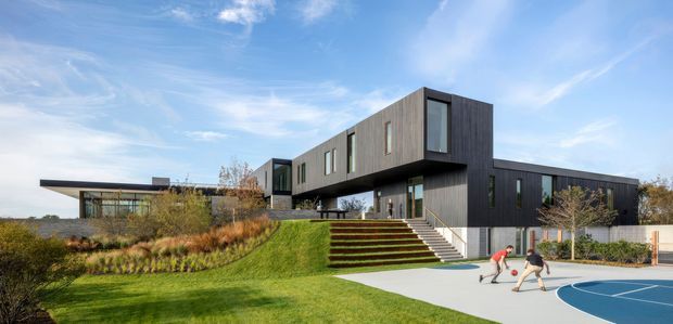 Exterior view of a contemporary multi-level building on a landscaped sloping site. The design features dark vertical timber cladding, extensive glazing, strongly cantilevered box-like volumes, a broad low roofline, and a stepped entry path leading toward an outdoor basketball court.