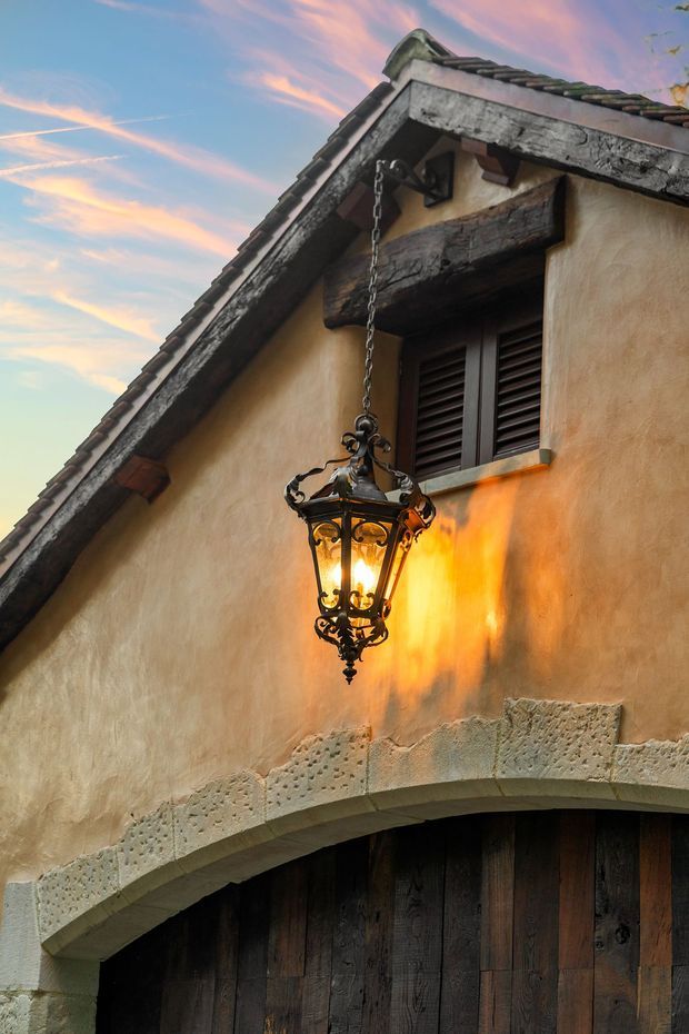 Close-up exterior view of a rustic, traditional stucco building facade at dusk. The steep gable roof has dark clay-like roof tiles and heavy weathered timber trim and brackets. A wrought-iron hanging lantern illuminates the warm plaster wall above a large dark timber arched garage or entry door, with a small louvred timber window beneath the gable.