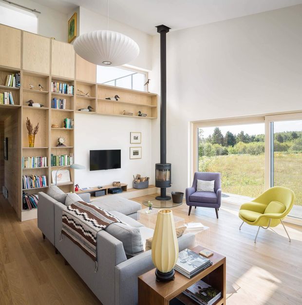 Bright, airy living room with extensive built-in light-wood shelving and cabinetry, a wall-mounted television, grey sectional sofa, colourful accent chairs, a central freestanding black stove, and large windows overlooking greenery. The room has light timber flooring, white walls, layered contemporary furnishings, and a sculptural pendant light.