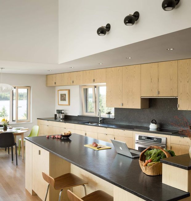 Bright open-plan kitchen with extensive light timber cabinetry, dark stone benchtops and splashback, a large central island with seating, and integrated cooking appliances. The kitchen opens to a dining area and large windows, with black spherical ceiling and wall-mounted lighting adding contrast.
