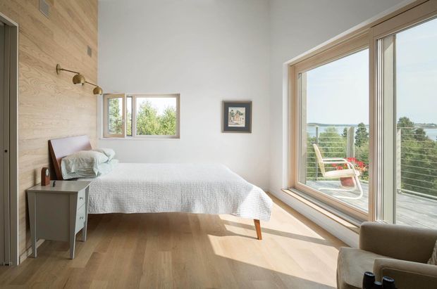 A bright, minimalist bedroom with a single bed, pale timber flooring, and a full-height glazed corner opening onto a balcony or terrace. The room features a warm timber feature wall with simple brass reading lights, white walls, a small bedside table, and a calm view toward water and greenery outside.
