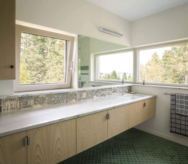 Bright mid-century-inspired bathroom with a long wall-mounted timber vanity, white countertop, double sinks, large corner windows, and a decorative patterned tile backsplash. The room has a green tiled floor, pale neutral walls, and a wall-mounted towel radiator, with trees visible outside.