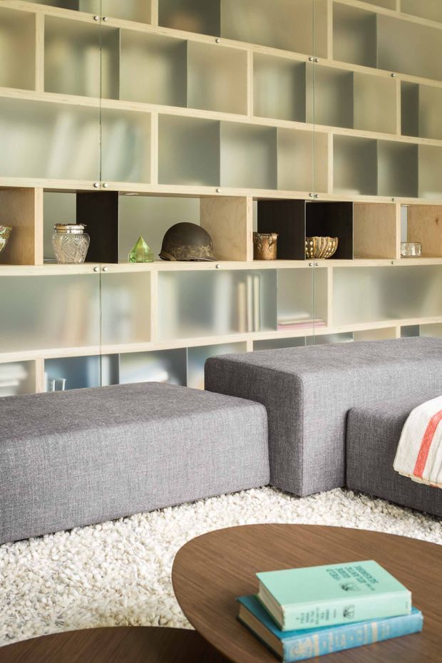A contemporary living room featuring a modular grey upholstered sectional sofa, a low rounded wooden coffee table with books, and a white textured rug. The main architectural feature is a large wall-to-wall built-in shelving system with pale timber framing, translucent or frosted sliding panels, and open display niches containing decorative objects. The space is softly lit with a neutral, refined palette.