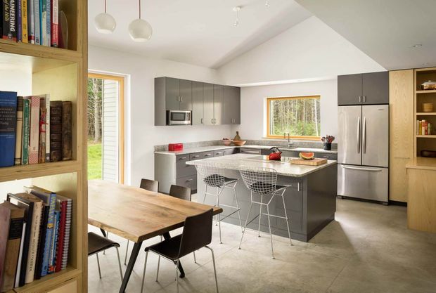 Open-plan contemporary kitchen and dining area with a central island, integrated stainless-steel appliances, dark grey cabinetry, light timber shelving and joinery, and a timber dining table. Large windows and a glazed door provide views to greenery and bring in natural light. Wire-frame bar stools and simple pendant lights add a modern, functional character.
