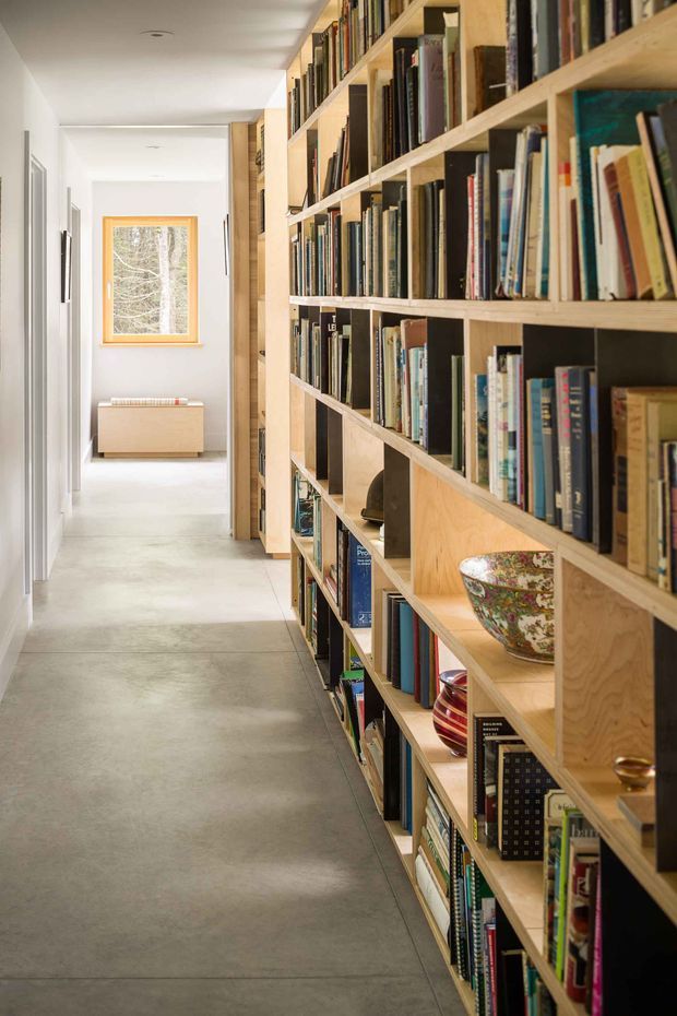 A bright, narrow hallway lined on the right with extensive floor-to-ceiling open timber shelving filled with books and decorative objects. The corridor has smooth white walls and ceiling, a pale polished concrete floor, simple recessed ceiling lights, and a timber-framed window at the far end above a low radiator or cabinet. A doorway opens along the left wall, creating a clean, gallery-like circulation space.