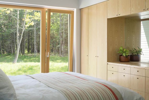 A bright, minimalist bedroom with a low bed in the foreground, floor-to-ceiling timber-framed glazing opening toward a wooded garden, and a full-height built-in timber wardrobe and dresser wall. The joinery includes a recessed display niche with potted plants, while soft white bedding and pale timber create a calm, natural atmosphere.