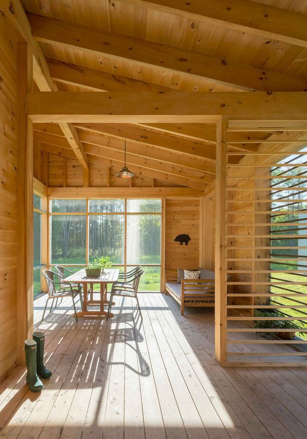 A warm, timber-lined enclosed porch or sunroom with exposed structural beams and a pitched timber ceiling. Floor-to-ceiling glazing opens views to a wooded landscape, while a slatted timber screen divides the space. The room contains a small dining table with chairs, a simple timber bench, pendant lighting, and rustic outdoor footwear, creating a bright natural retreat.