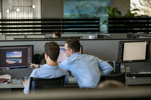 A contemporary open-plan office with two coworkers collaborating at a workstation. Multiple computer monitors, shared desks, dark horizontal privacy screens, pendant lighting, and a large abstract artwork are visible. The space has a professional, collaborative atmosphere with muted grey, black, and blue tones.