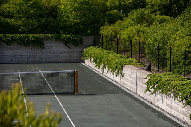 A landscaped outdoor tennis court with dark playing surface and white boundary lines, enclosed by low concrete retaining walls topped with black mesh fencing. Cascading greenery and mature trees surround the court, creating a private garden setting.