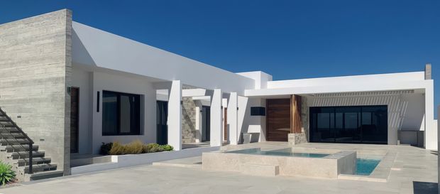 Exterior view of a low-profile contemporary residence arranged around a paved courtyard and raised rectangular pool. The white rendered structure has strong geometric volumes, a flat roofline, expansive dark-framed glazing, a textured concrete feature wall, timber entry cladding, exposed stone accents, and exterior stairs.