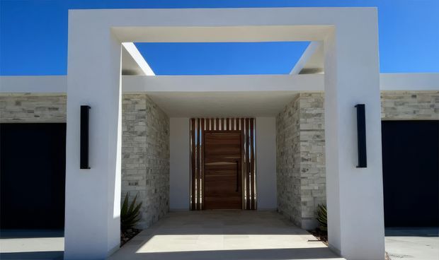 Front entry facade of a contemporary residence, featuring a white rendered rectangular portal with an open skylit canopy, pale stacked-stone walls, a warm timber pivot door with vertical slatted screening, and dark garage doors on either side. Strong sunlight and deep shadows create a crisp, high-contrast exterior composition.