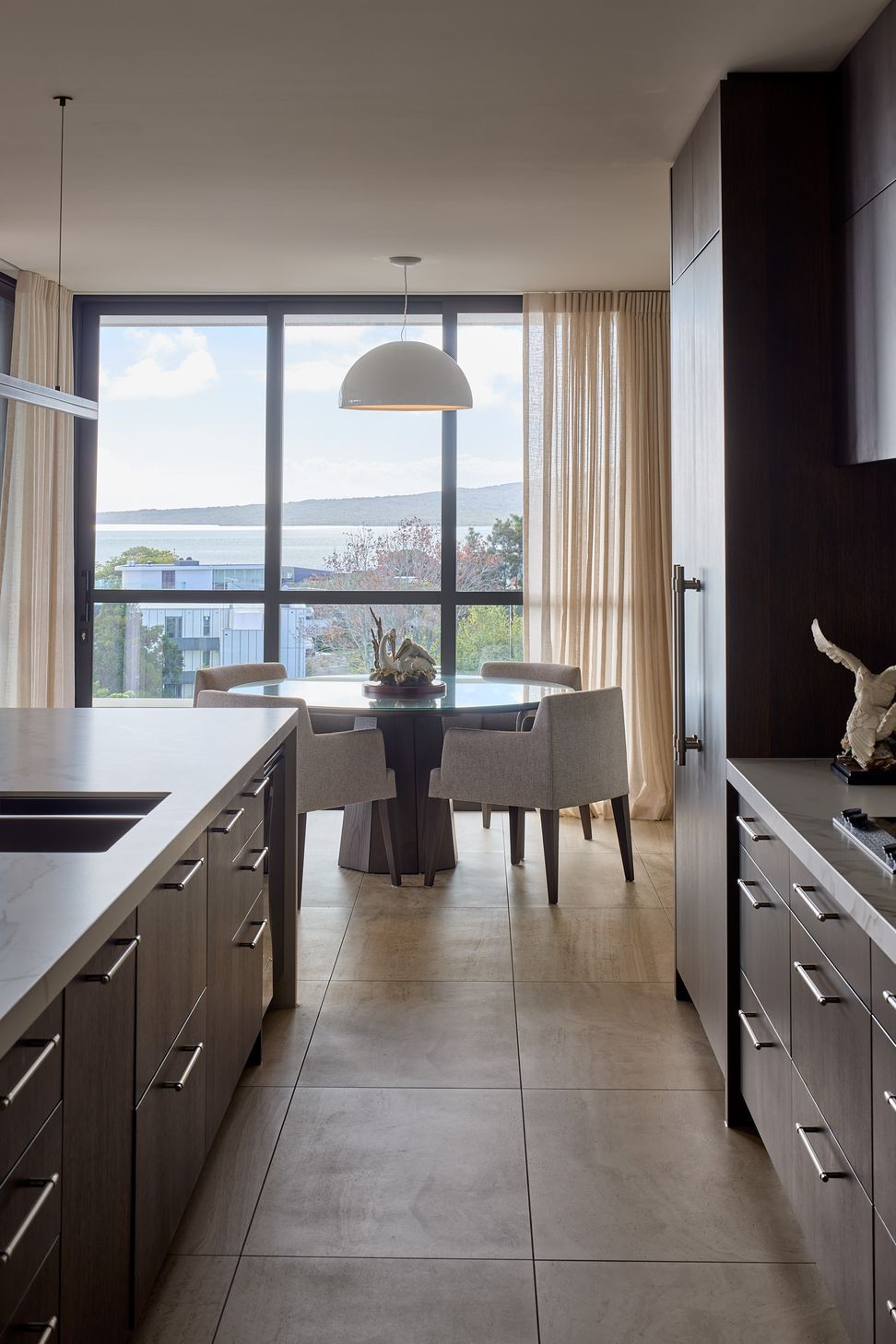 From the kitchen, views extend beyond the informal round dining area towards the Hauraki Gulf. Dark cabinetry, pale stone benchtops and tiled flooring create a sophisticated and enduring interior palette.