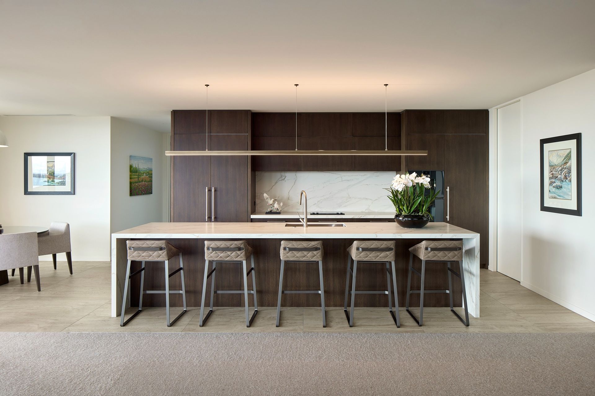 The generous kitchen island provides seating for five and forms the social centre of the open-plan living space. Pale stone surfaces contrast with dark bespoke cabinetry, while a linear pendant emphasises the island's length and the concealed scullery maintains a calm, uncluttered interior.