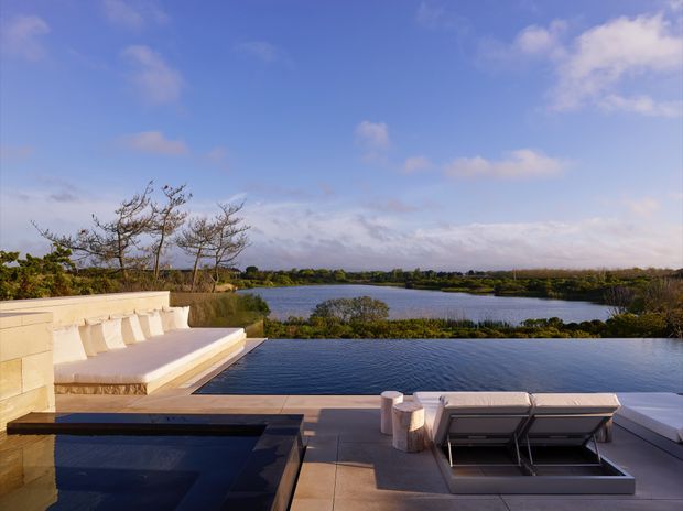 A spacious rooftop or elevated outdoor terrace overlooking a calm body of water and landscaped wetlands. The scene includes a dark rectangular pool or water feature in the foreground, pale stone paving and built-in bench seating, and contemporary outdoor lounge furniture. The setting is open, serene, and strongly connected to the waterfront landscape under a blue, lightly clouded sky.