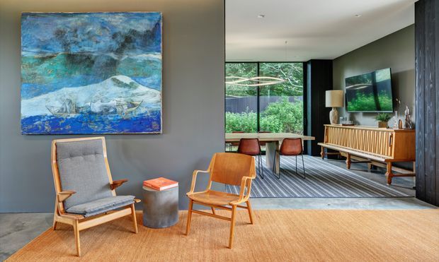 A stylish mid-century-inspired living room with warm timber furniture, grey upholstered lounge chairs, a cylindrical side table, and a large abstract blue artwork. The room opens into a dining and media area with a timber credenza, television, pendant lighting, and expansive glazing overlooking greenery. Dark vertical wall panelling, pale walls, timber flooring, and a large neutral rug create a refined, warm interior.