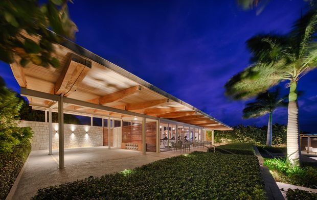 Night-time exterior view of a low, single-storey modern building with an extensive timber-lined roof overhang supported by slender posts. The facade combines warm timber, large glazed panels and pale masonry walls, with illuminated covered outdoor areas, manicured hedges and palm trees around the building.
