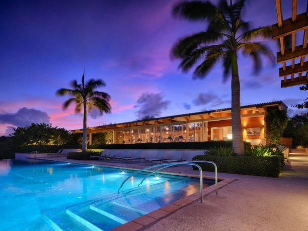 Twilight exterior view of a contemporary single-storey residence with extensive glazed living spaces, warm timber-toned illumination, a large illuminated swimming pool with steps and stainless-steel handrails, and tall palm trees. The home has a broad low-pitched roof and an open covered terrace overlooking the pool.