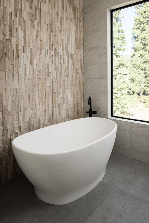 A modern bathroom featuring a freestanding white oval bathtub beside a large black-framed window. The walls combine a vertically textured light timber-look feature surface with large pale grey stone-look tiles, while the floor is finished in grey tile. A matte black wall-mounted bath tap provides contrast, and trees are visible outside the window.