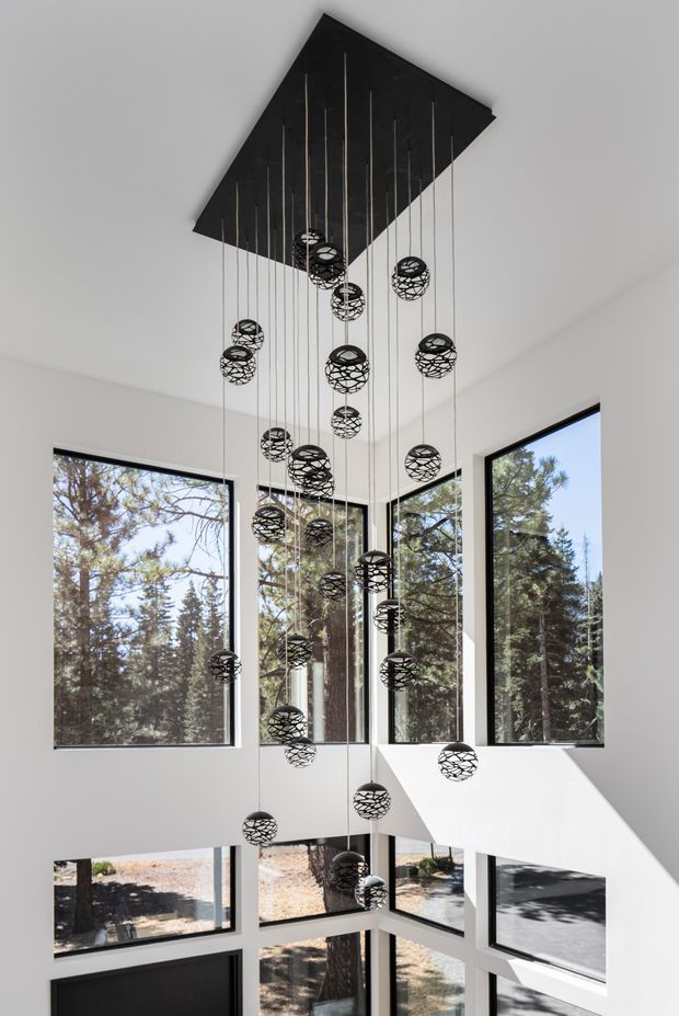 A dramatic double-height interior void with extensive floor-to-ceiling corner glazing overlooking trees. A large black rectangular ceiling canopy supports numerous suspended spherical pendant lights at varying heights. White walls, black window frames, and strong natural daylight create a modern architectural composition.