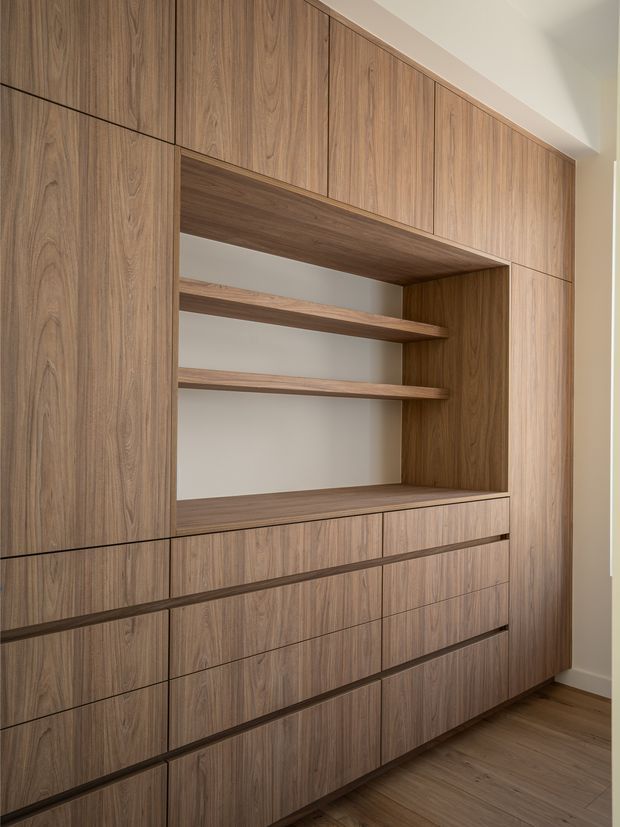 Built-in floor-to-ceiling timber-toned cabinetry with integrated drawers, tall doors, and an open recessed display niche with floating shelves. The joinery has a clean, handleless design and is set against a light neutral wall with matching wood flooring.