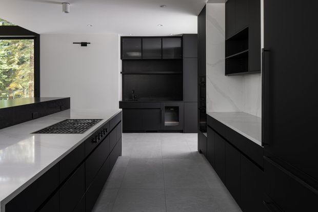 A sleek contemporary kitchen viewed from the main work area, with extensive matte-black cabinetry, a large island with integrated gas cooktop, pale stone benchtops, grey large-format tiled flooring, and a dark built-in wall unit at the rear. White walls and ceiling provide contrast, while a large black-framed window brings in views of greenery.