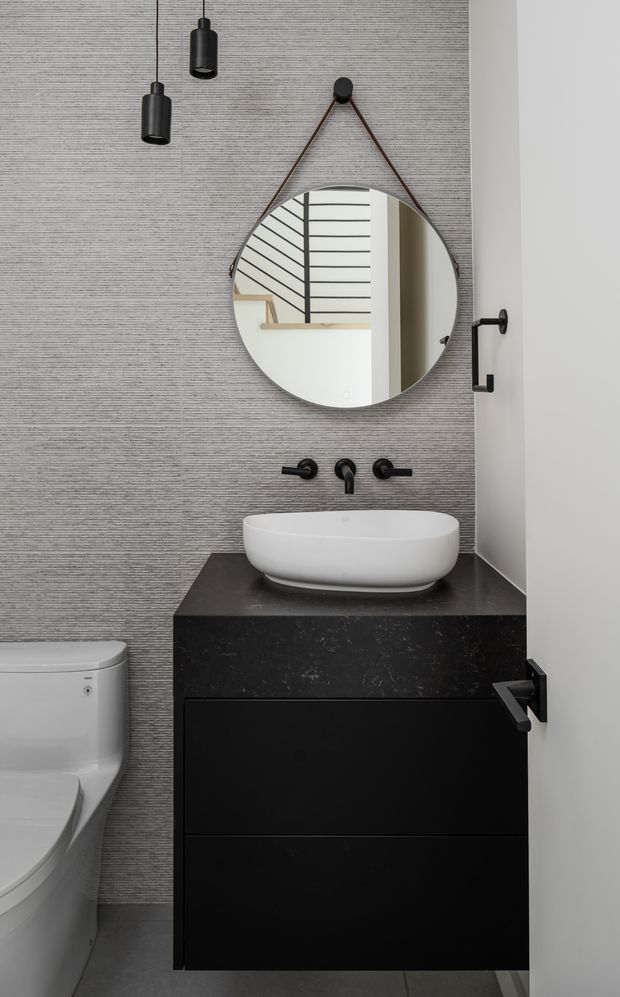 A compact contemporary bathroom featuring a floating black vanity with integrated dark stone benchtop, a white vessel basin, matte black wall-mounted tapware, and a round hanging mirror. The grey vertically textured wall finish, black pendant lights, and concealed toilet create a refined monochromatic scheme.