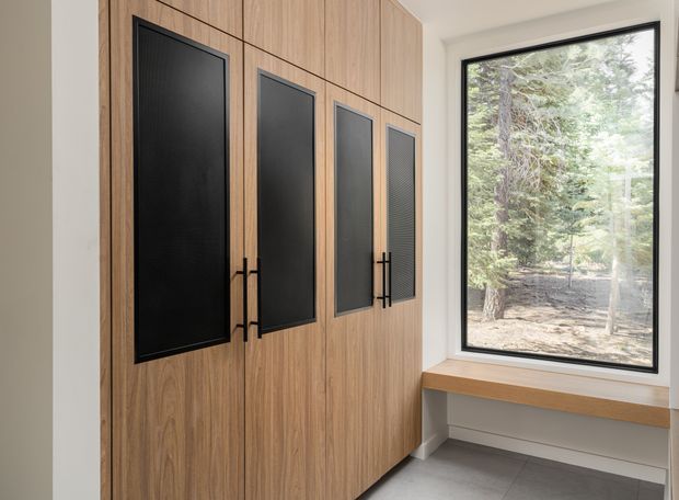 A contemporary built-in storage wall with warm wood-grain cabinetry and tall black inset panel fronts, slim black handles, and integrated dark grille-like panels. Beside it is a large black-framed picture window overlooking trees, with a timber window seat or ledge below. The space has light painted walls and large grey floor tiles, creating a clean, refined, nature-connected interior.