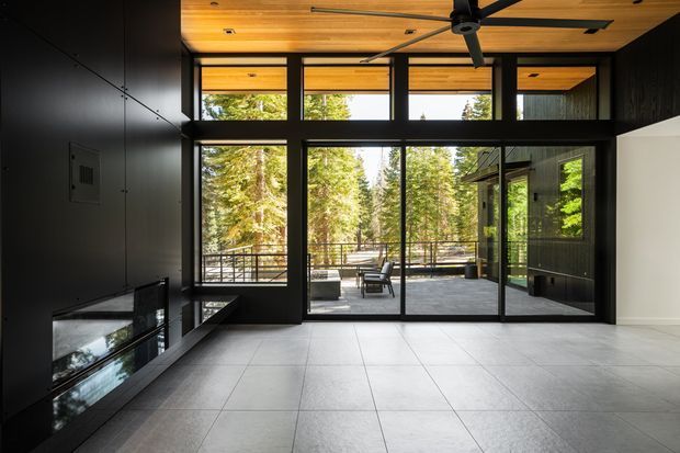 A dramatic contemporary living room or media room with dark charcoal built-in cabinetry and wall panels, expansive black-framed floor-to-ceiling glazing, and a warm timber-lined ceiling with an exposed ceiling fan. Pale large-format tiled flooring contrasts with the dark interior. The glazing opens to a covered outdoor terrace surrounded by tall forest trees, creating a strong indoor-outdoor connection. A low built-in media unit is visible along the left wall.