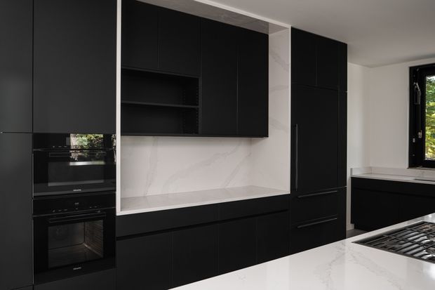 A contemporary black kitchen with full-height matte cabinetry, integrated double ovens, open shelving, a large white marble-look splashback and benchtop, and a gas cooktop in the foreground. A window brings in natural light from the side.