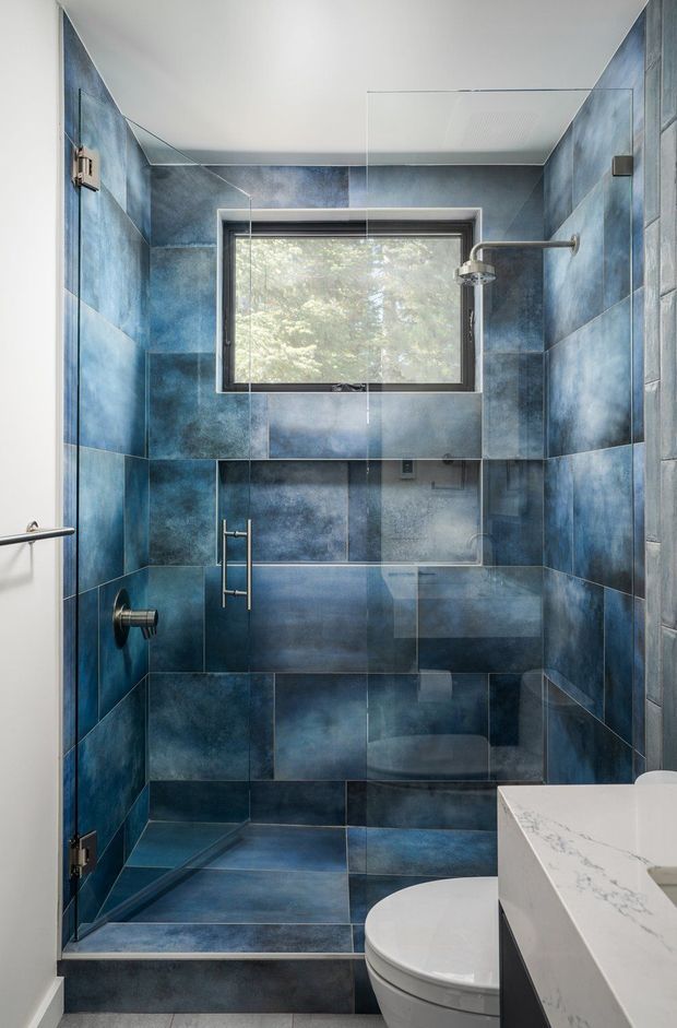 A compact contemporary bathroom featuring a walk-in glass shower enclosed by vivid blue, cloud-patterned wall and floor tiles. The shower includes a rain showerhead, handheld fixture, glass screen and built-in niche, with a black-framed window providing natural light and views of trees. A white toilet and a white marble-look vanity with dark cabinetry are visible in the foreground.