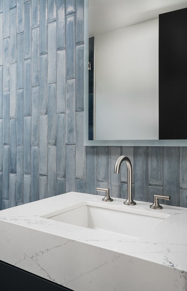 Close-up of a refined bathroom vanity featuring a white engineered-stone benchtop with an integrated rectangular basin, brushed silver gooseneck tapware, dark cabinetry, a large frameless mirror, and vertically arranged blue-grey textured wall tiles.