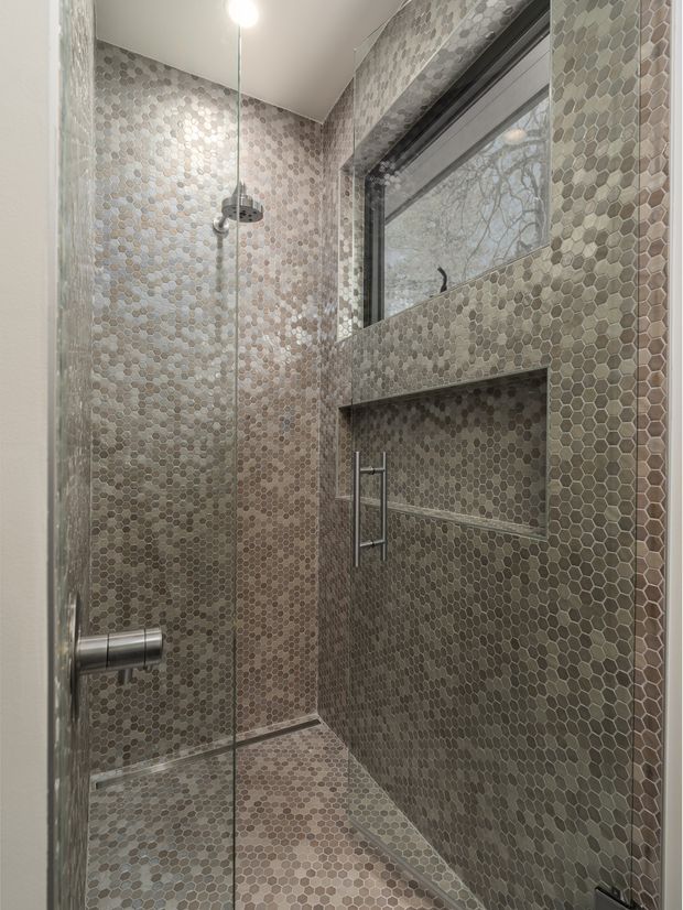 Compact contemporary walk-in shower enclosed by clear glass, with small hexagonal mosaic tiles covering the walls, floor, recessed shelf, and window surround. A chrome rain shower, handheld fixture, frameless glass door, and horizontal window create a clean, highly tiled bathroom interior.