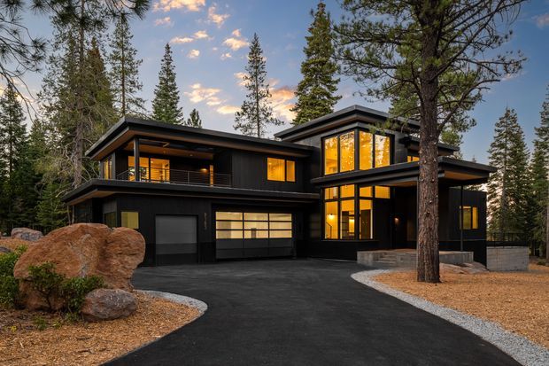 Exterior view of a contemporary multi-level dark-clad house set among mature pine trees. The home features broad flat roof planes, extensive warm-lit glazing, projecting upper volumes, a balcony, and an asphalt driveway curving around landscaped rock and gravel areas.