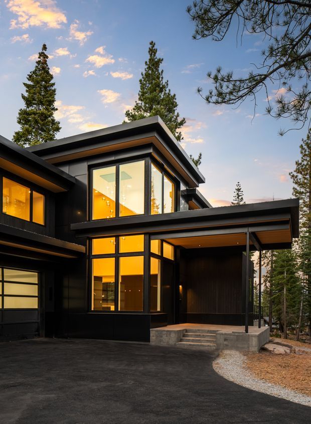 Exterior view of a contemporary multi-level house set among tall pine trees. The building has a dark black-clad facade, extensive floor-to-ceiling glazing glowing warmly from inside, strong horizontal roof overhangs, a flat/low-pitched roofline, and a covered entry porch with concrete steps. A dark paved driveway and gravel border occupy the foreground.