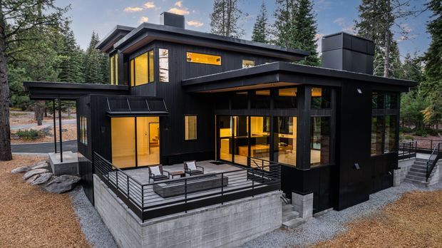 A contemporary multi-level black-clad house set among tall forest trees. The building has a combination of flat and low-pitched roof forms, extensive warm-lit glazing, a raised concrete foundation, and a large outdoor terrace with black metal balustrades and outdoor seating. Exterior stairs and gravel landscaping surround the home.